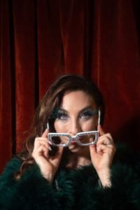 Jess looking straight the camera putting on a pair of flamboyant, Elton John style glasses